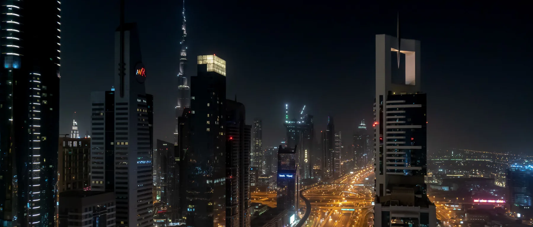 Die Sheikh Zayed Road bei Nacht mit den beleuchteten Türmen der Downtown Dubai und dem Burj Khalifa in der Ferne