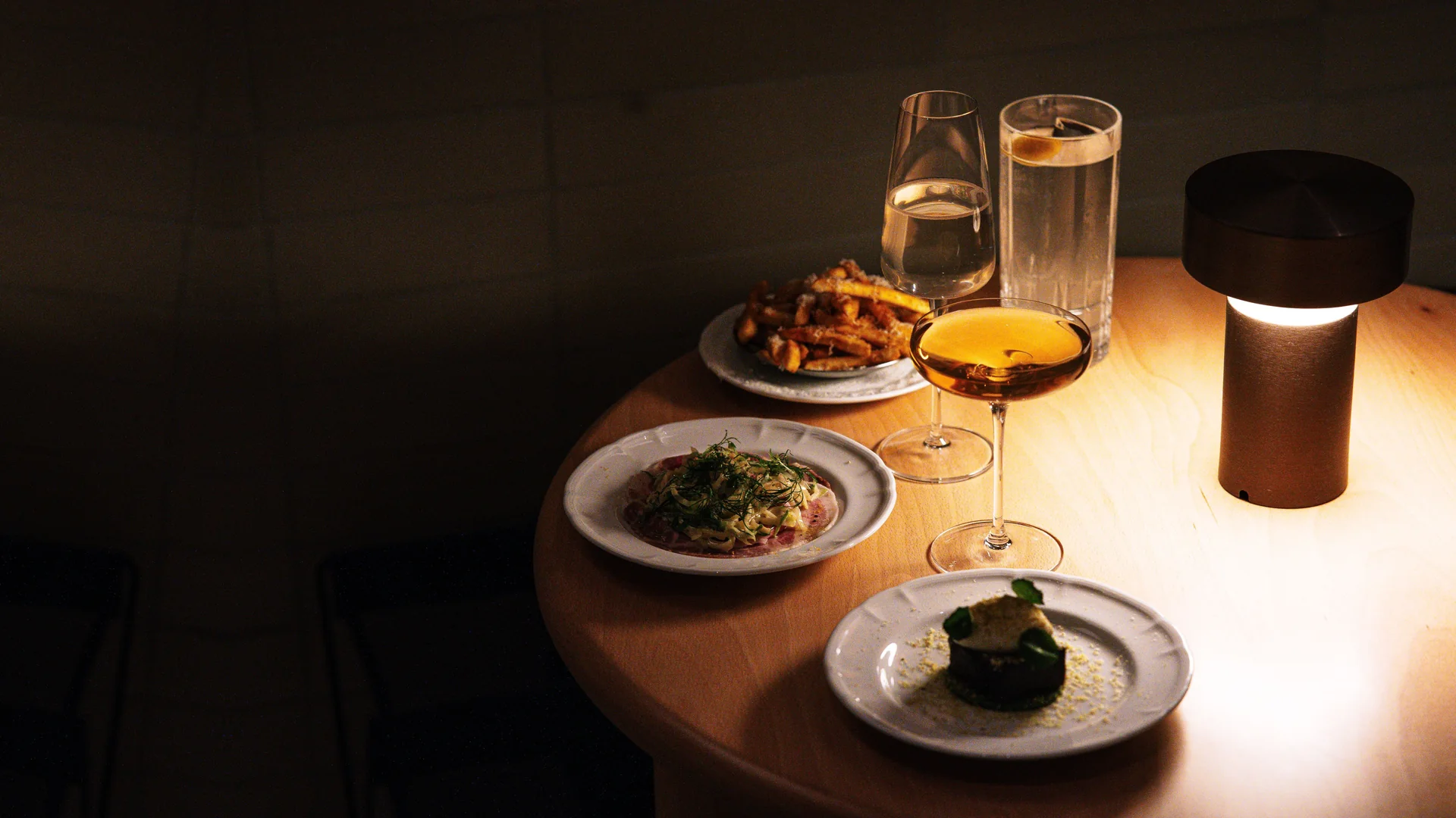 Ein kleiner runder Tisch in einem dunklen Raum, beleuchtet von einer einzigen Lampe, gedeckt mit angerichteten Speisen und einer Coupe bernsteinfarbenen Cocktails