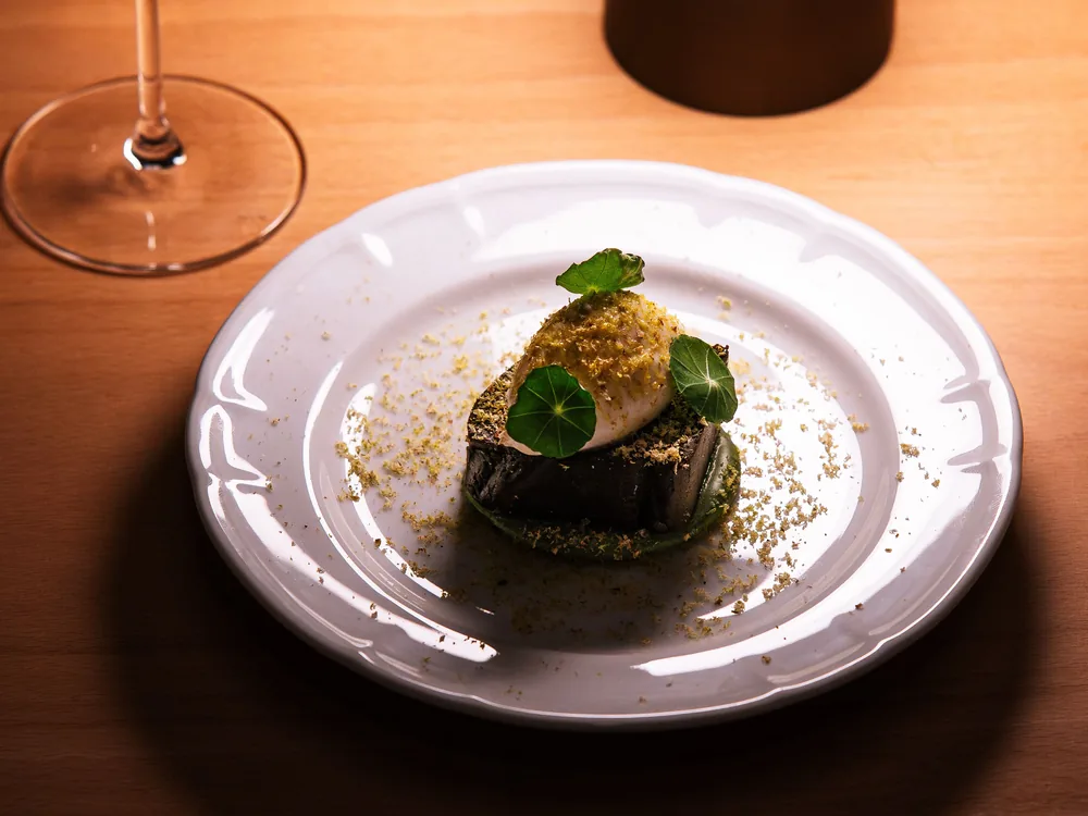 Ein einzelnes angerichtetes Dessert mit Kapuzinerkressenblättern auf einem weißen Teller mit Wellenrand, warm beleuchtet auf einem Holztisch
